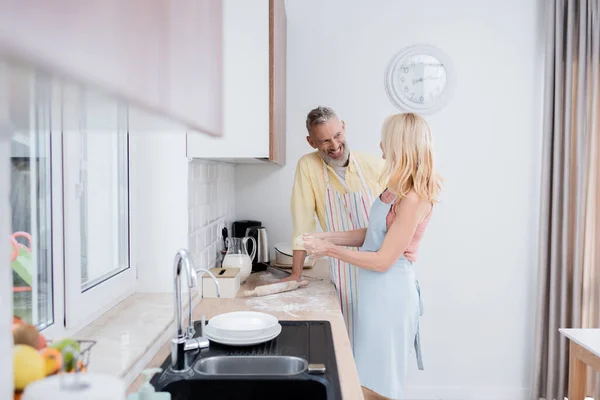 Sourire homme regardant femme faire de la pâte près de la farine et rouleau à pâtisserie dans la cuisine — Photo de stock