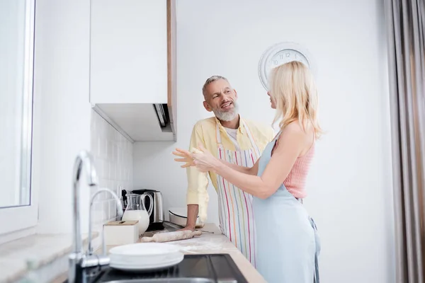 Позитивний зрілий чоловік розмовляє з дружиною з тістом на кухні — стокове фото