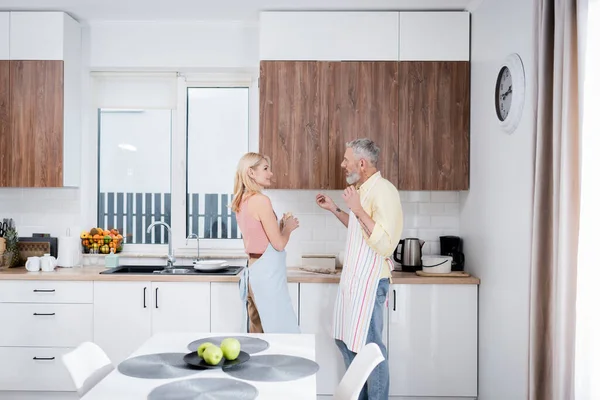 Side view of mature couple talking while cooking dough in kitchen — Stock Photo