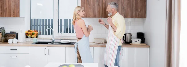 Vista lateral del hombre maduro hablando con la esposa cocinar masa en la cocina, pancarta - foto de stock