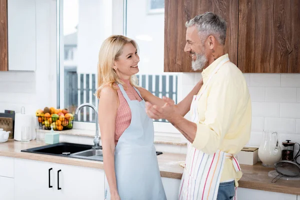 Sonriendo hombre maduro en delantal hablando con la esposa en la cocina - foto de stock