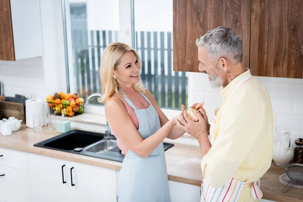 Femme blonde souriante dans un tablier tenant la pâte près du mari dans la cuisine — Photo de stock