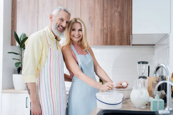 Hombre positivo en delantal mirando a la cámara cerca de la esposa y los ingredientes en la cocina - foto de stock