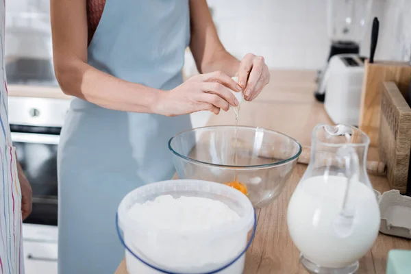 Vista recortada de la mujer vertiendo huevo en un tazón cerca de los ingredientes y el marido en la cocina — Stock Photo