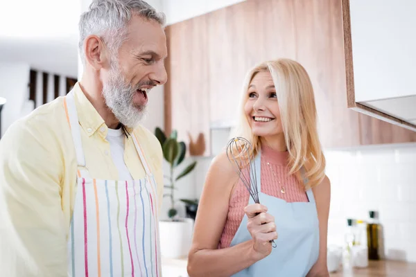 Fröhliche Frau in Schürze hält Schneebesen neben reifem Mann in Küche — Stockfoto