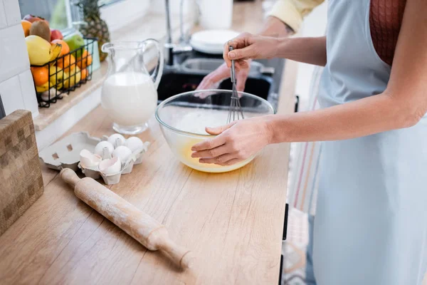 Vista ritagliata di donna in grembiule mescolando uova in ciotola vicino mattarello e marito a casa — Foto stock