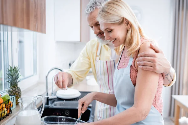 Мужчина наливает муку и обнимает жену венчиком на кухне — стоковое фото