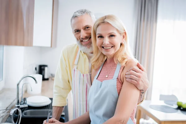 Alegre pareja madura en delantales mirando a la cámara en la cocina - foto de stock