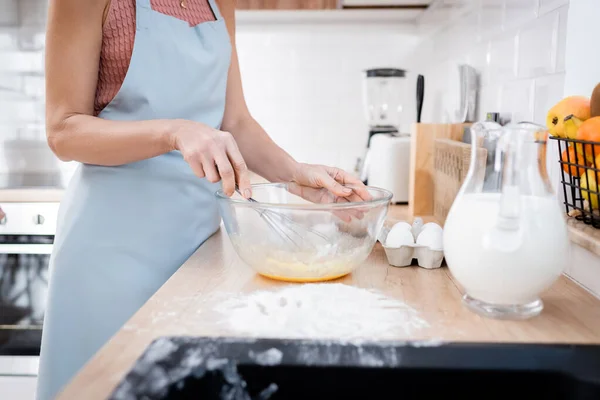 Vue recadrée de la femme dans un tablier mélangeant des œufs dans un bol près de la farine et du lait sur le plan de travail de la cuisine — Photo de stock