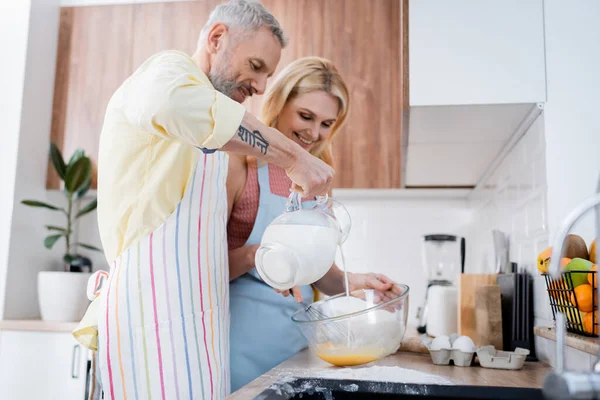 Smiling man pouring milk in bowl near wife cooking in kitchen — Stock Photo