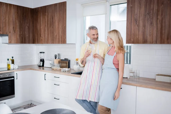 Sorridente coppia matura in grembiuli che tengono champagne in cucina — Foto stock