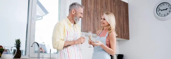Mature woman in apron smiling at husband with champagne in kitchen, banner — Stock Photo
