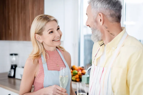 Cheerful mature woman holding glass of champagne and talking to blurred husband in kitchen — Stock Photo