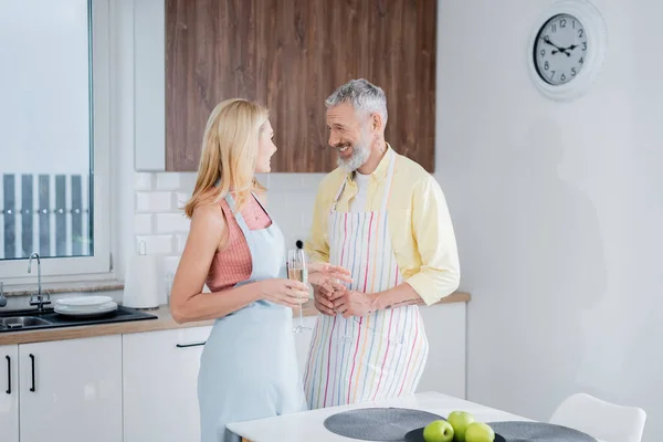 Gai mature homme tenant champagne près de femme dans tablier dans cuisine — Photo de stock