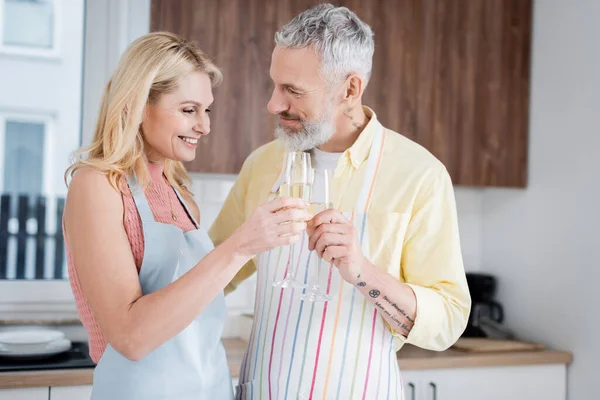 Sonriente mujer madura tintineo champán con marido en delantal en casa - foto de stock