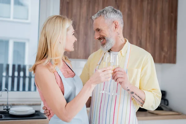 Vista laterale di sorridente uomo di mezza età in grembiule clinking champagne con moglie a casa — Foto stock