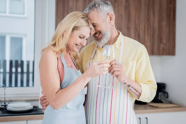 Feliz pareja madura en delantales tintineo champán en casa - foto de stock