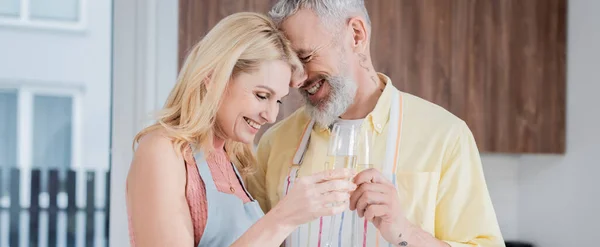 Femme mûre souriante cliquetis champagne avec mari dans tablier à la maison, bannière — Photo de stock