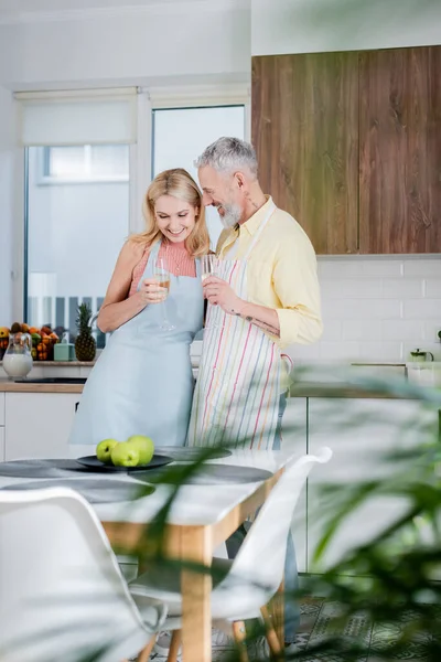 Позитивный зрелый мужчина в фартуке держит шампанское рядом с женой в фартуке на кухне — стоковое фото