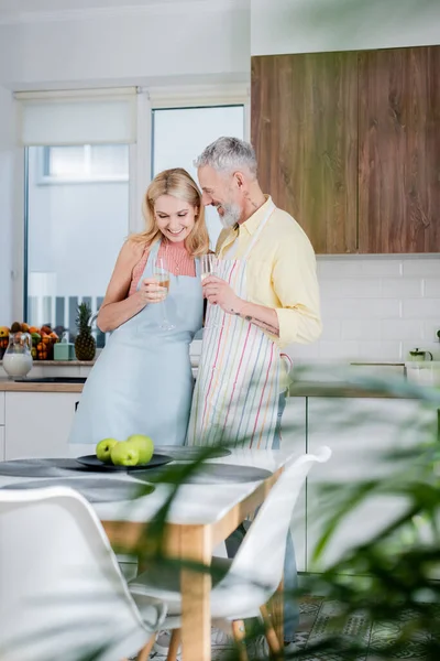 Positivo uomo maturo in grembiule azienda champagne vicino moglie in grembiule in cucina — Foto stock