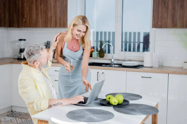 Mujer sonriente en delantal sosteniendo champán cerca del marido utilizando el ordenador portátil en la cocina - foto de stock