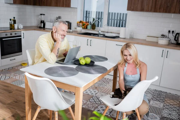 Uomo sorridente guardando moglie vicino al gatto e laptop a casa — Foto stock