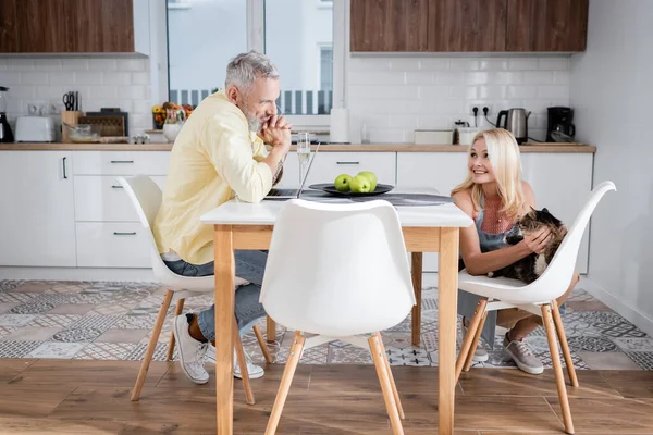 Positive woman petting cat and looking at husband near laptop and champagne at home — Stock Photo