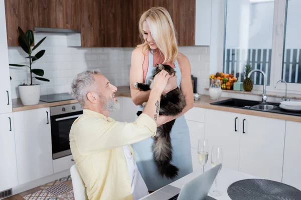 Positiva donna matura in grembiule con gatto vicino marito, laptop e champagne a casa — Foto stock