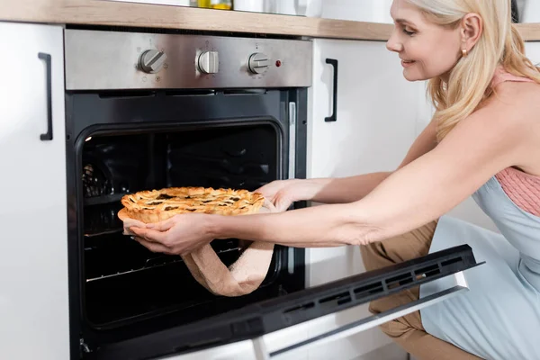 Vista lateral da mulher madura positiva que toma torta do forno na cozinha — Fotografia de Stock