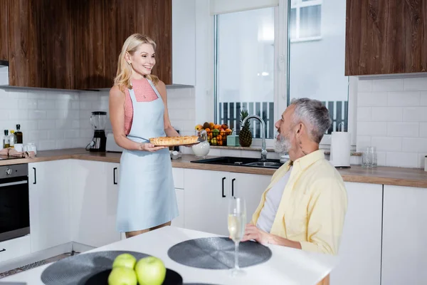 Positive Frau hält hausgemachten Kuchen neben Ehemann und Champagner auf Tisch in Küche — Stockfoto