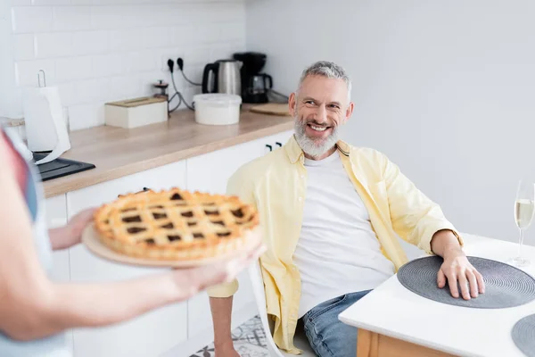 Positive man looking at blurred wife with pie near champagne in kitchen — Stock Photo