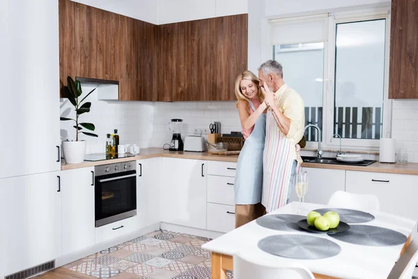 Homme mûr dansant avec femme souriante dans tablier près de champagne dans la cuisine — Photo de stock