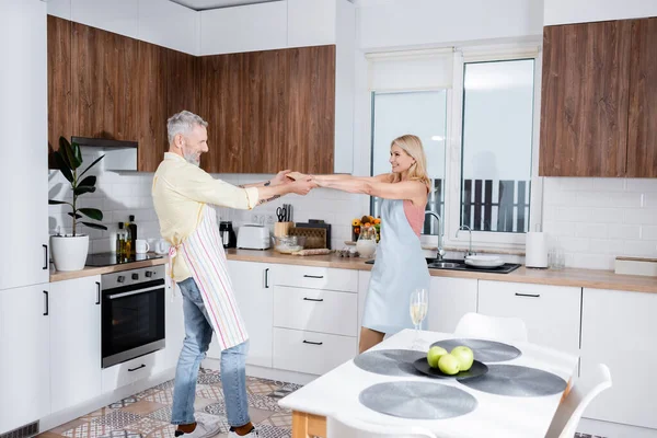 Casal positivo em aventais dançando perto de champanhe na mesa na cozinha — Fotografia de Stock