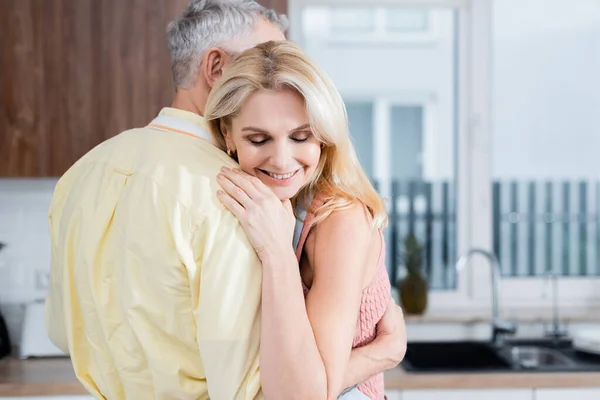 Donna sorridente con gli occhi chiusi che abbraccia il marito in cucina — Foto stock