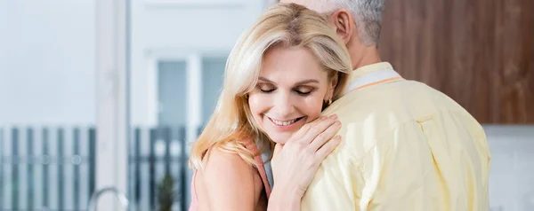 Mujer madura sonriendo mientras abraza marido en la cocina, pancarta - foto de stock