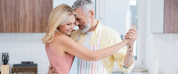 Positiv reifer Mann in Schürze tanzt mit lächelnder Frau in Küche, Banner — Stockfoto