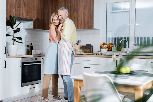 Alegre pareja madura en delantales abrazándose cerca de champán en la cocina - foto de stock