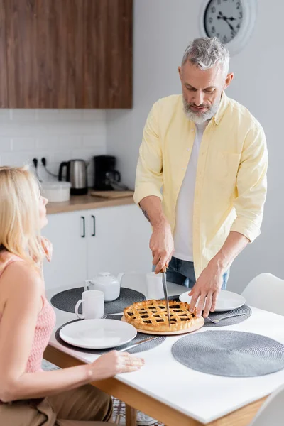 Maduro hombre corte casero pastel cerca borrosa esposa en cocina - foto de stock