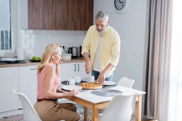 Мужчина режет пирог рядом с улыбающейся женой и чаем на кухне — стоковое фото