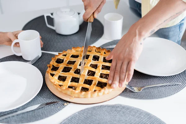 Vista recortada del hombre cortando pastel cerca de tazas y esposa en la cocina - foto de stock