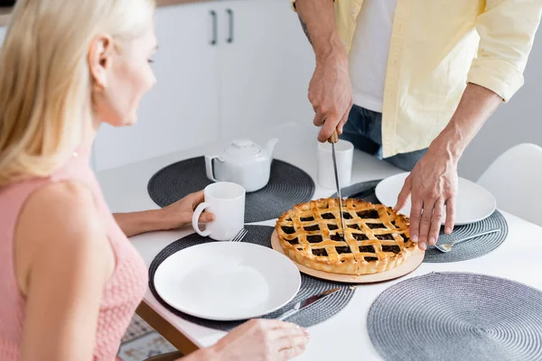 Mature man cutting pie near blurred wife with tea at home — Stock Photo