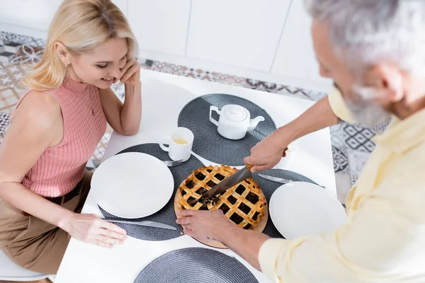 Вид сверху на размытого человека, резающего домашний пирог рядом с улыбающейся женой на кухне — стоковое фото