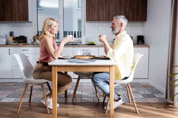 Vista laterale di coppia matura positiva che tiene tazze vicino a torta in cucina — Foto stock