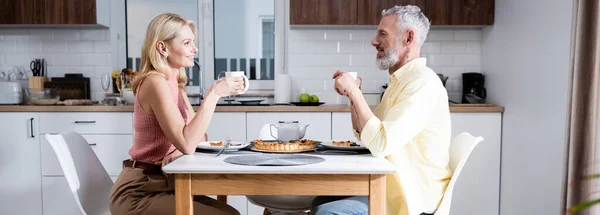 Vista lateral de la mujer sonriente sosteniendo la taza cerca del marido y el pastel en la cocina, pancarta - foto de stock