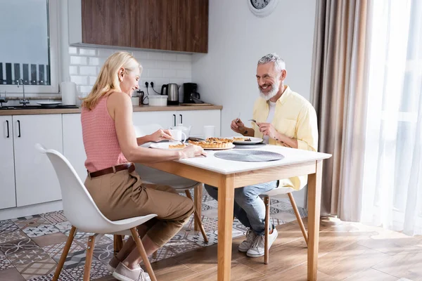 Fröhlich reif mann holding besteck in der nähe von ehefrau und kuchen im küche — Stockfoto