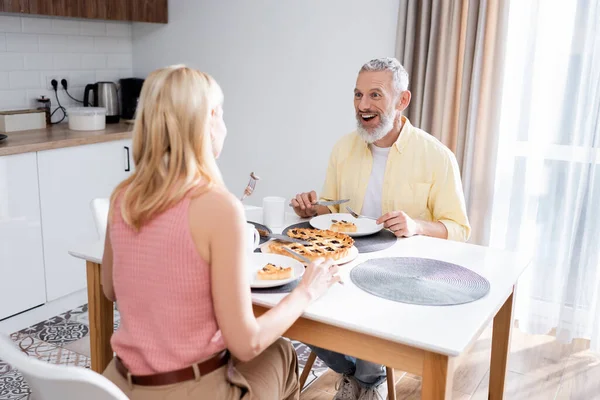 Emocionado hombre sosteniendo cubiertos cerca borrosa esposa y pastel en la cocina - foto de stock