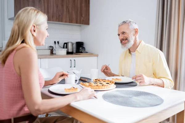 Hombre maduro sosteniendo cubiertos cerca de pastel mientras habla con su esposa en la cocina - foto de stock