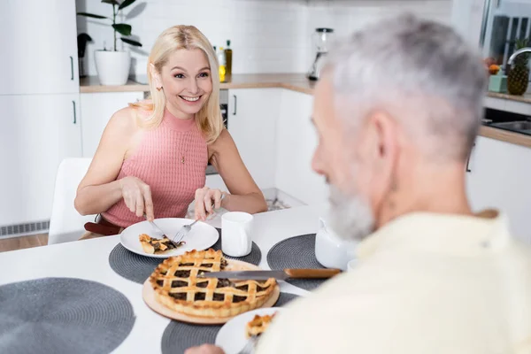 Mujer sonriente cortando pastel cerca de marido borroso en la cocina - foto de stock