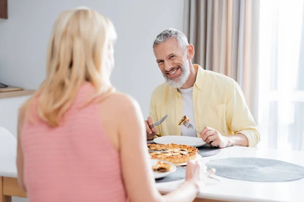 Positivo maduro hombre mirando borrosa esposa cerca de pastel en cocina - foto de stock
