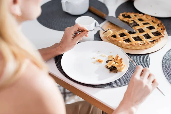 Vista ritagliata di donna offuscata che tiene posate vicino a torta saporita e tazza in cucina — Foto stock